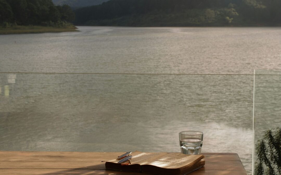 Open boek, leesbril en glas water op een houten tafel met uitzicht over een meer